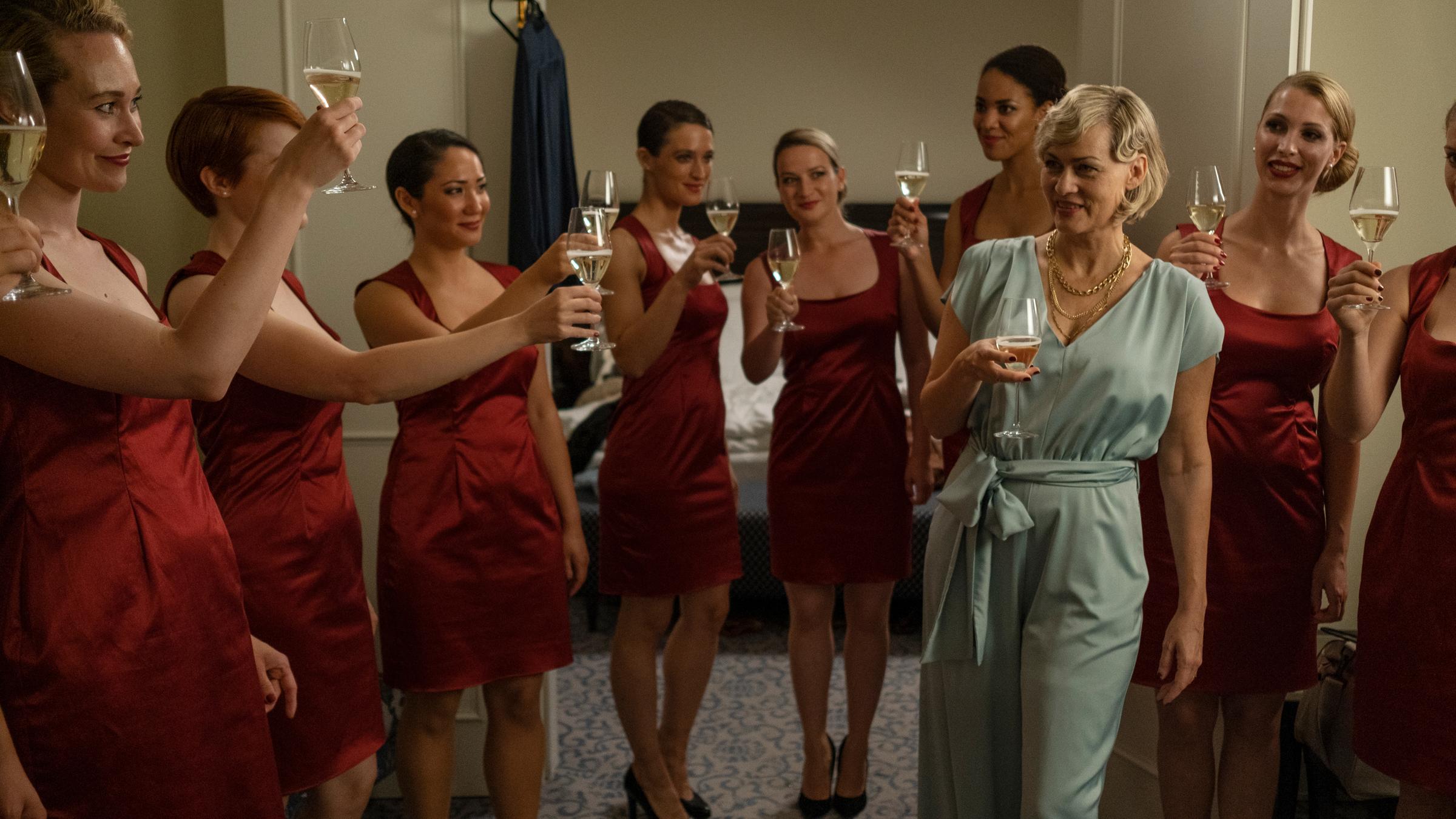 "So laut du kannst": Agenturchefin Ines Bezold (Gesine Cukrowski) und ihre Hostessen, darunter Kim Krawitz (Friederike Becht) und Maja Heller (Nina Gummich), stehen zusammen in roten Cocktailkleidern und prosten ihrer Vorgesetzen im eleganten Hosenanzug in einer Hotelsuite zu.