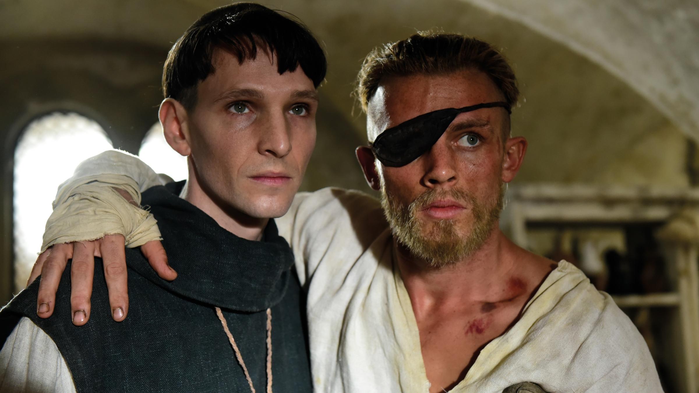 "Narziss und Goldmund": Nahaufnahme von Narziss (Sabin Tambrea) und Goldmund (Jannis Niewöhner), die in einem Gewölbe stehen. Narziss stützt den verletzten Goldmund, dessen Auge mit einer Augenklappe bedeckt ist.