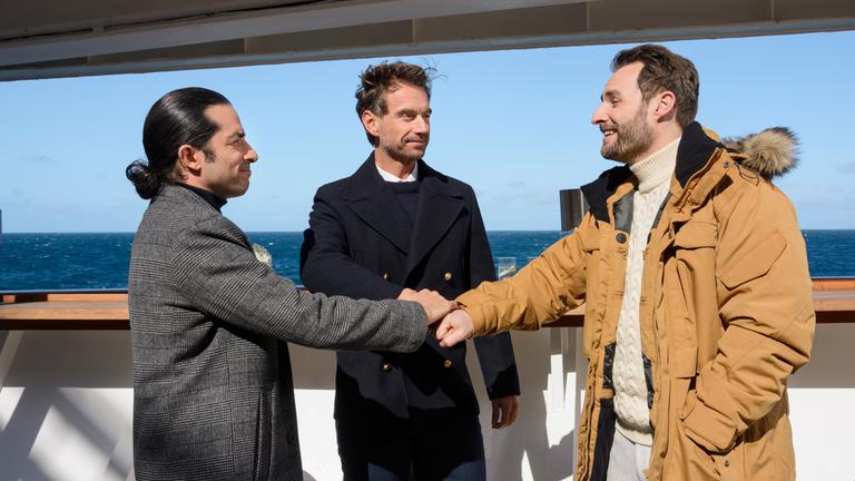"Das Traumschiff -  Lappland": An Deck des Schiffs stehen Max Parger (Florian Silbereisen), Patrick Steiner (Daniel Fritz) und Maik Berghammer (Tayfun Baydar) zusammen. Die Freunde legen die Hände übereinander und lächeln sich an.