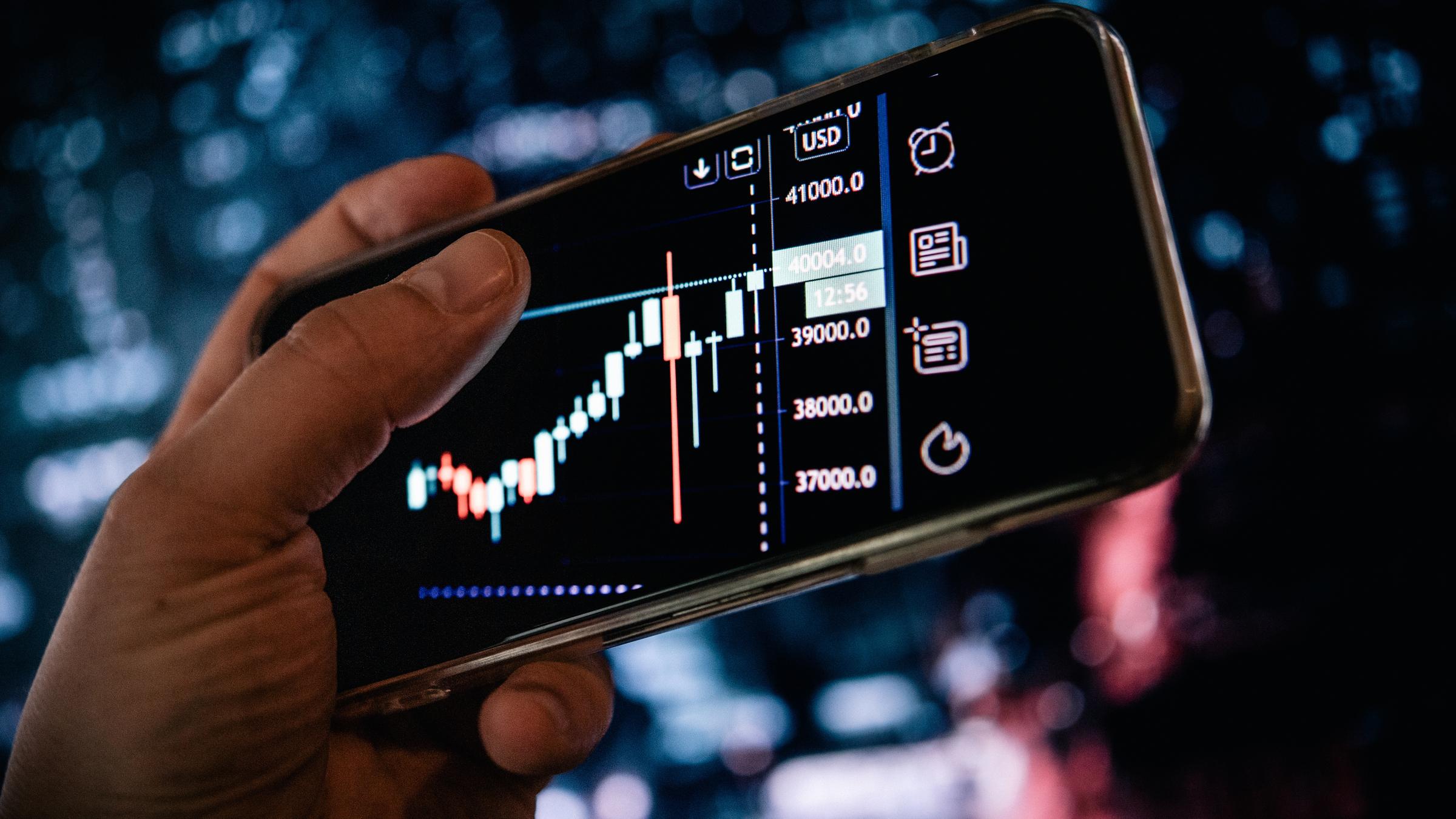 "WISO-Dokumentationen: Krieg, Kollaps, Kursverlust. Was die Krise mit unserem Geld macht": Eine Hand hält ein Handy, auf dem ein Kursverlauf zu sehen ist.