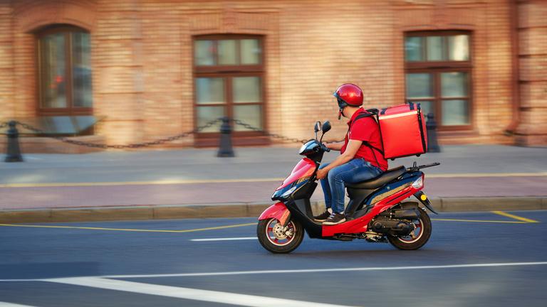 "Liefern am Limit - Die harte Welt der Bringdienste": Ein Lieferdienstfahrer fährt mit seinem Moped und seiner roten Tragetasche auf dem Rücken durch die Straßen.