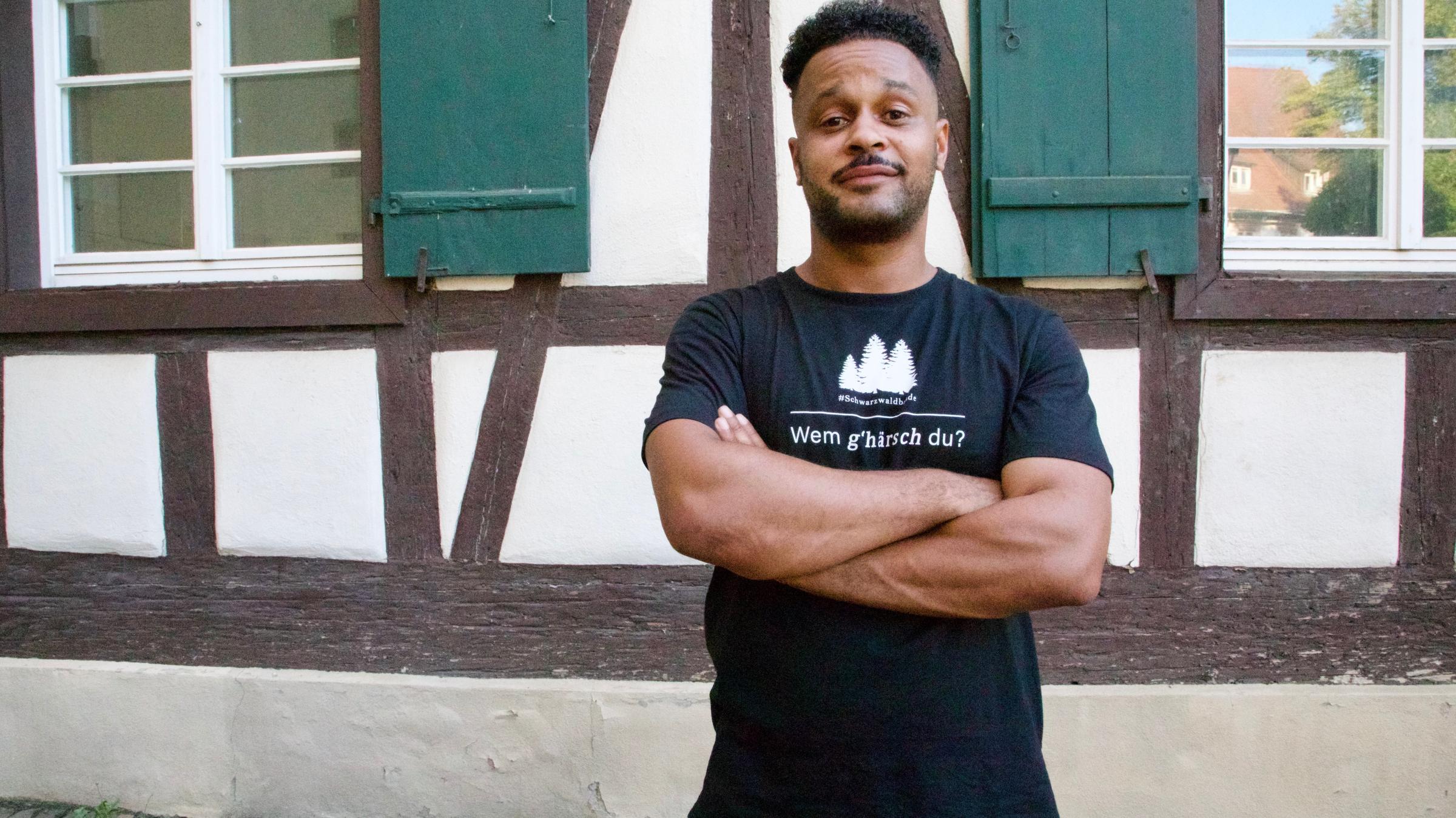"Ich bin deutsch": Lukas Steier alias Cossu steht mit verschränkten Armen vor der Wand eines Fachwerkhauses mit zwei Fenstern und grünen Klappläden.