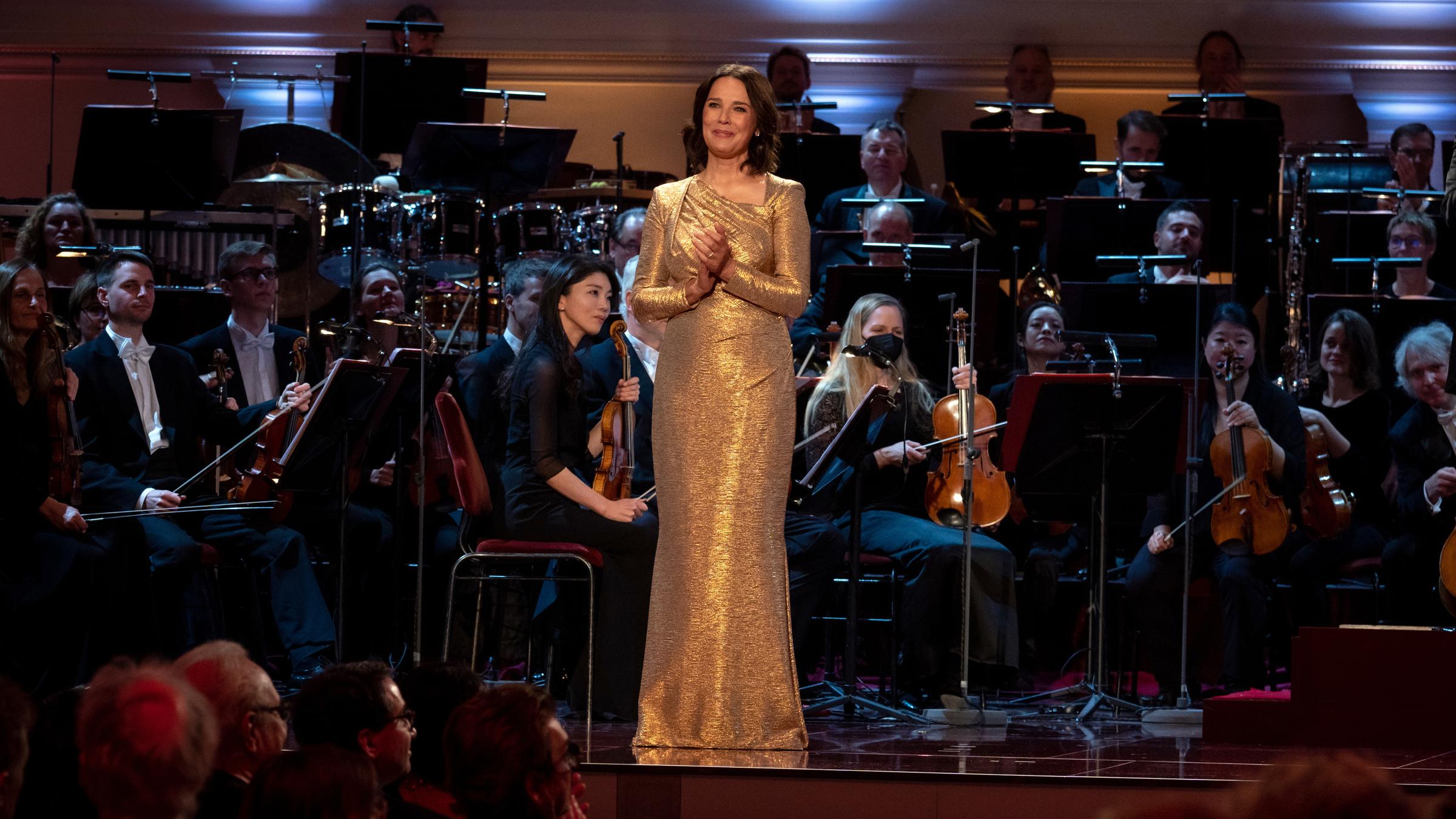 "OPUS KLASSIK 2022": Désirée Nosbusch steht auf einer Bühne. Sie hat die Hände vor dem Körper gefaltet und lächelt. Gekleidet ist sie mit einem goldfarbenen Abendkleid. Hinter ihr sitzen die Musiker*innen des Orchesters.