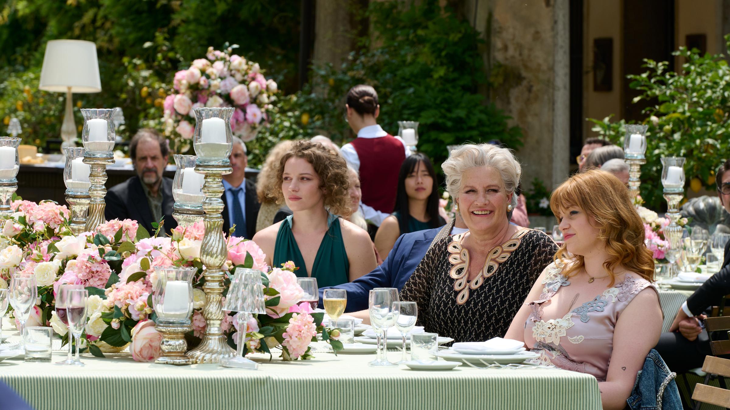 "Süßer Rausch - Die Familie": An einem festlich gedeckten Tisch mit riesigen  Blumenbouquets und Kerzenleuchtern sitzt Constanze (Suzanne von Borsody) zwischen Marguerite (Antonia Bill) und Simone (Lilly Charlotte Dreesen). Im Hintergrund beobachtet der Journalist Gerlich (Helmfried von Lüttichau) die Szene.