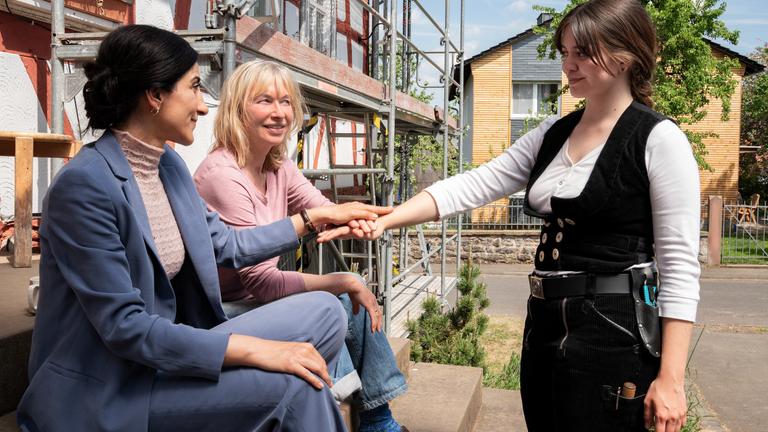 "Unterm Apfelbaum: Einsturzgefährdet": Jasmin (Saman Giraud) und Frieda (Therese Hämer) sitzen auf den Stufen vor dem Eingang von "Heckemellersch", einem Fachwerkhaus. Tinka (Lotte Becker) steht ihnen gegenüber. Alle drei legen zum Schwur die Hände in der Mitte übereinander.