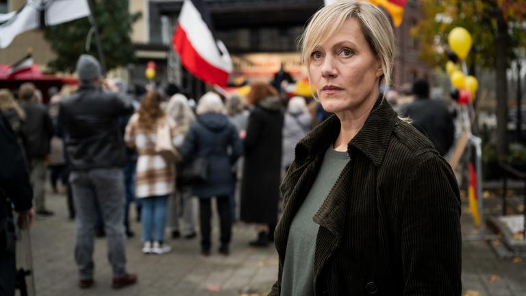 "Die Bürgermeisterin": Claudia Voss (Anna Schudt) steht am Rande eines öffentlichen Platzes, auf dem sich mehrere Menschen, einige mit Fahnen, versammelt haben, und schaut nachdenklich um sich.