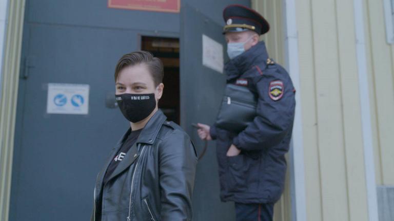  "Russlands mutige Frauen - Widerstand gegen Putin": Zwei Personen stehen im Halbprofil vor der Tür zu einem Gebäude. Links eine junge Frau mit kurzem Haar, rechts ein uniformierter Polizist, der den Türgriff festhält. Beide tragen einen Mundschutz.