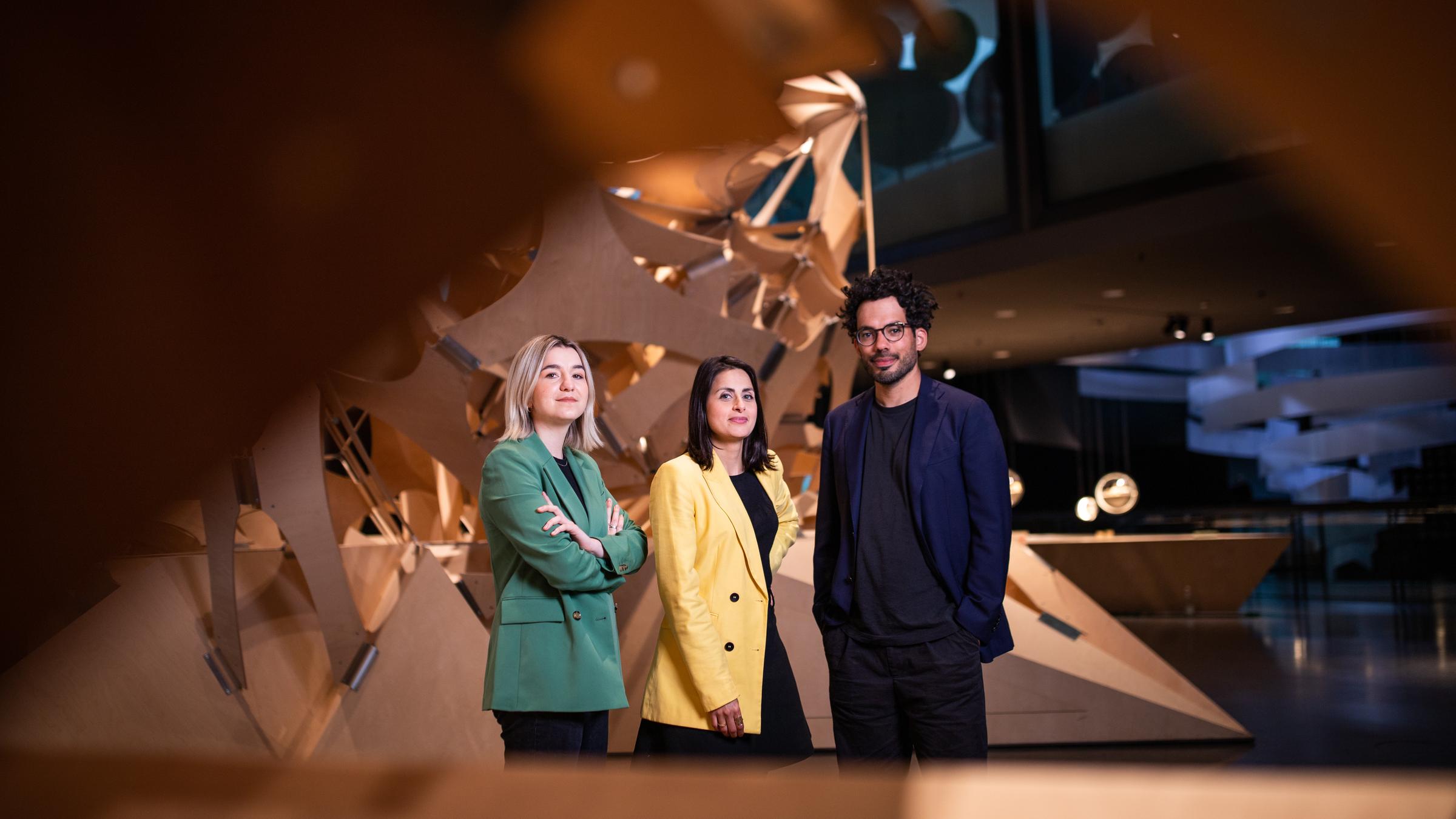 "Futur Wir – Das neue deutsche Selbstverständnis (1/3) - Wer wir sein wollen": Die drei Moderatoren stehen im Berliner Futurium und blicken in die Kamera. Eingerahmt sind sie von einer großen Holzskulptur.