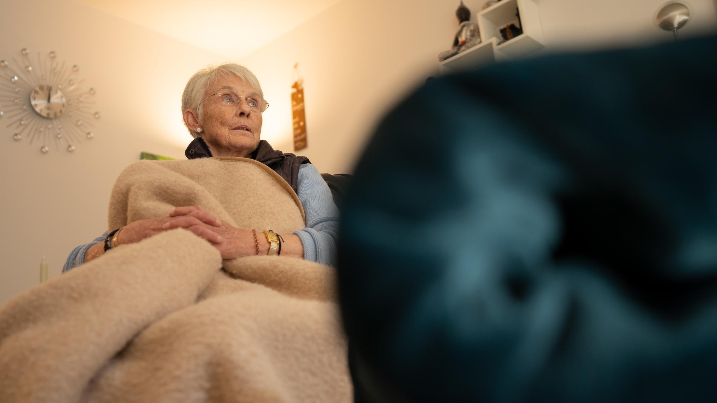 "ZDF.reportage: Wenn das Geld nicht reicht - zum Heizen": Eine Seniorin sitzt in einem Sessel und hat sich in eine Wolldecke gewickelt. Im Hintergrund hängt eine dekorative Wanduhr. Auf einem Wandregal sitzt eine Buddhafigur.