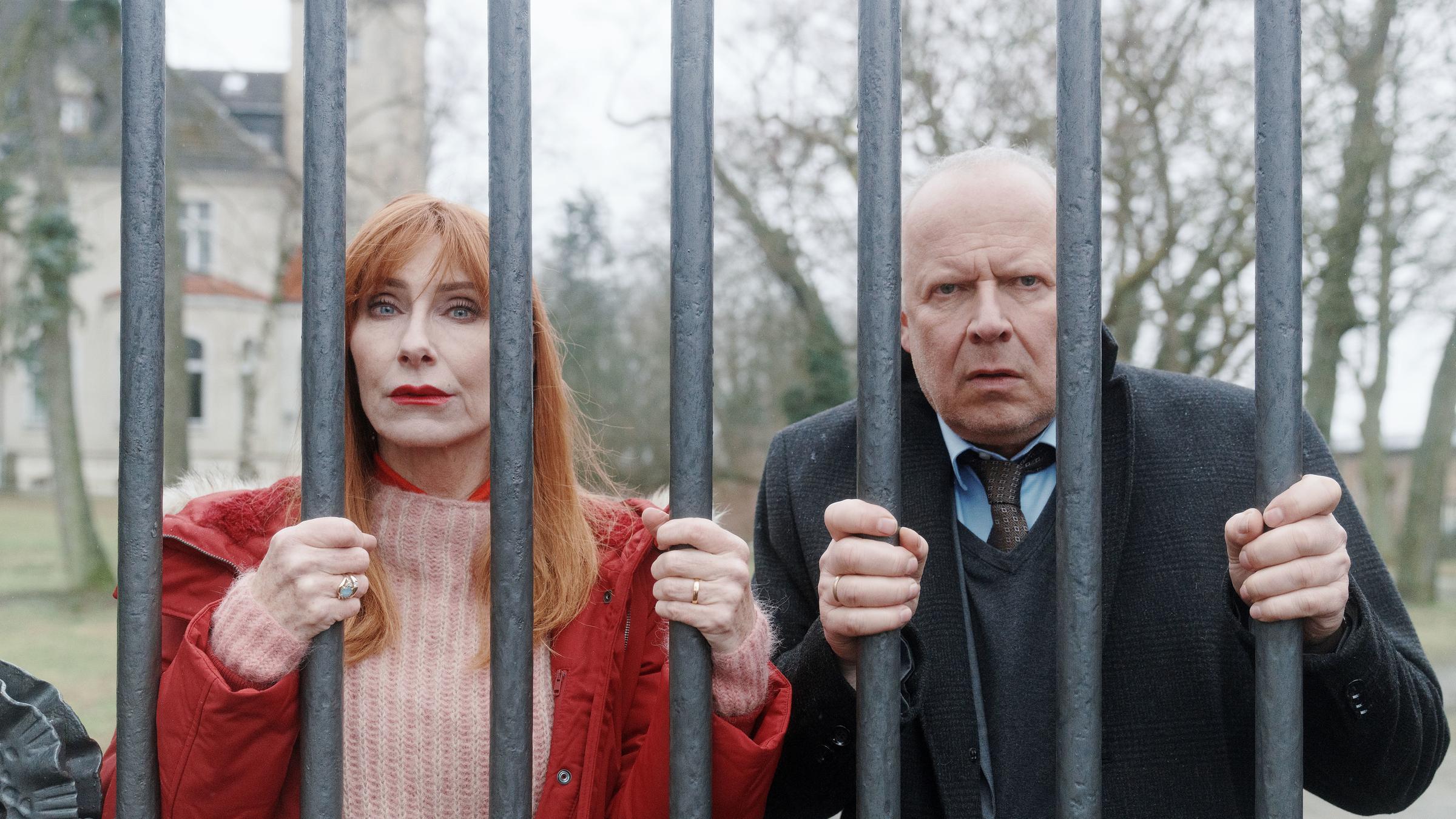 "Familie Bundschuh - Unter Verschluss": Gerald ( Axel Milberg) und Gundula (Andrea Sawatzki) krallen sich an ihrem Eisenzaun fest und blicken hilflos durch die dicken Gitterstäbe. Im Hintergrund ist der großangelegte Garten und ein Teil des Gutshauses zu sehen.