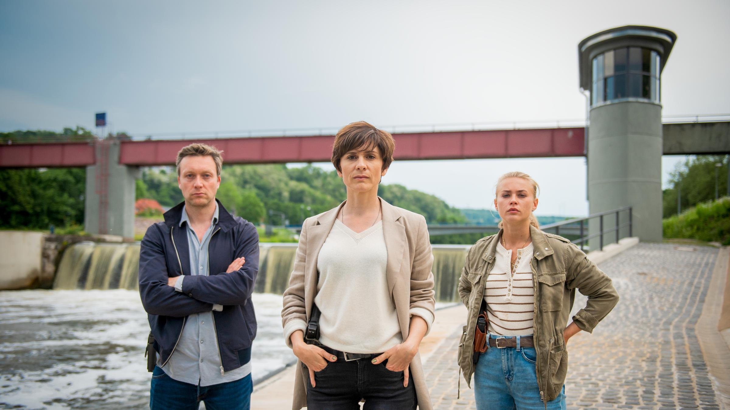 "In Wahrheit – Unter Wasser": Die Ermittler Freddy Breyer (Robin Sondermann), Judith Mohn (Christina Hecke), Lisa (Jeanne Goursaud) alle mit Kamerablick am Flussufer der Nied.