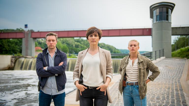 "In Wahrheit – Unter Wasser": Die Ermittler Freddy Breyer (Robin Sondermann), Judith Mohn (Christina Hecke), Lisa (Jeanne Goursaud) alle mit Kamerablick am Flussufer der Nied.