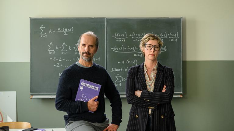 "Lehrer kann jeder!": Richard Glossat (Christoph Maria Herbst) sitzt in einem Klassenzimmer auf dem Lehrerpult und hält ein Mathematik-Buch in der Hand. Neben ihm steht Anke Glossat (Brigitte Zeh) mit verschränkten Armen und genervtem Blick. Im Hintergrund eine Schultafel, an die mit Kreide Mathematik-Formeln geschrieben stehen.