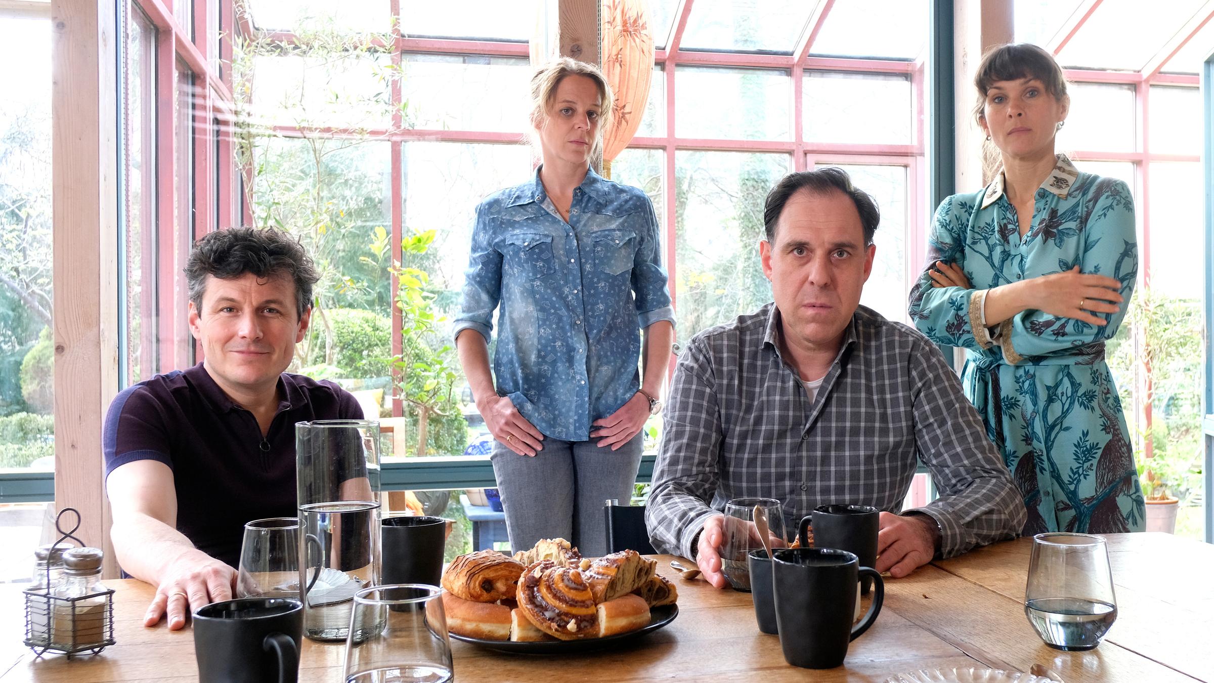 "Annie und das geteilte Glück": Nils (Manuel Rubey) und Ralf (Thomas Loibl) sitzen an einem Tisch, der mit Tassen, Gläsern und süßen Teilchen gedeckt ist, und schauen in die Kamera. In ihrem Rücken stehen Annie (Bernadette Heerwagen) und Tine (Kathrin von Steinburg) und schauen ebenfalls ernst in die Kamera.
