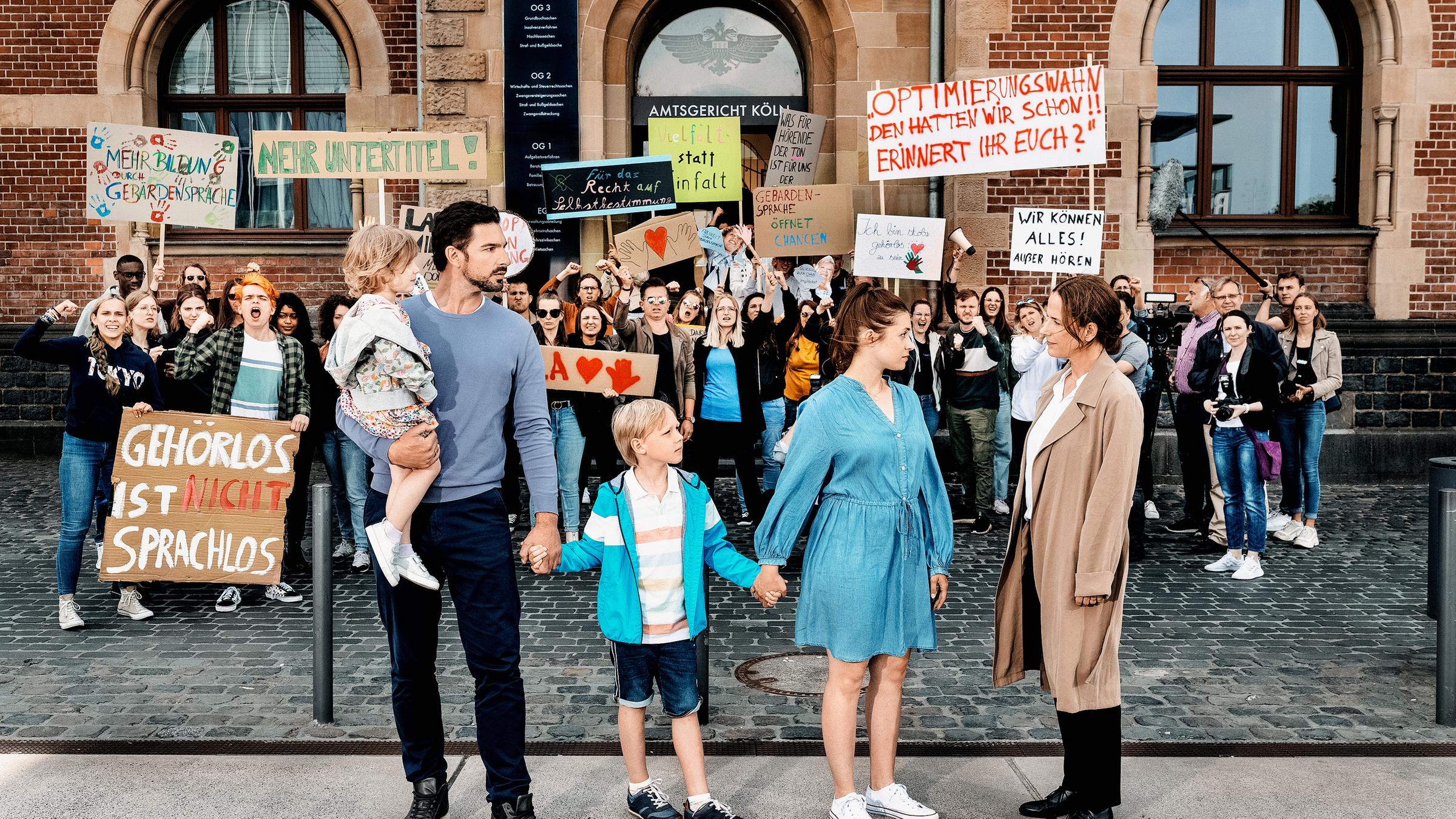 "Du sollst hören‘": Simon Ebert (Benjamin Piwko), Conny Ebert (Anne Zander), Mila Ebert (Delia Pfeffer), Mats Ebert (Leif-Eric Werk) und Jolanda Helbig (Claudia Michelsen) stehen vor dem Amtsgericht in Köln. Die Familie schaut zu Jolanda, Jolandas Blick fällt auf Conny Ebert. Hinter ihnen stehen Demonstranten.
