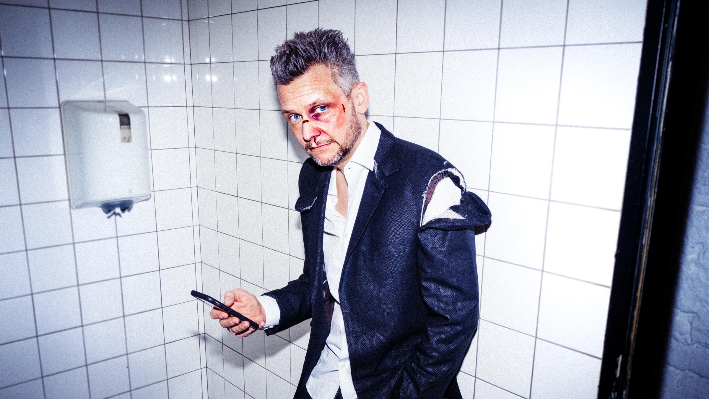 "Tobias Mann: Mann gegen Mann": Tobias Mann steht in einer öffentlichen Toilette vor dem Waschbecken, angelehnt an den Wandfliesen. Er sieht aus, als hätte er gerade eine Schlägerei gehabt, zerrissene  Klamotten, im Gesicht frische Wunden und ein blaues Auge.