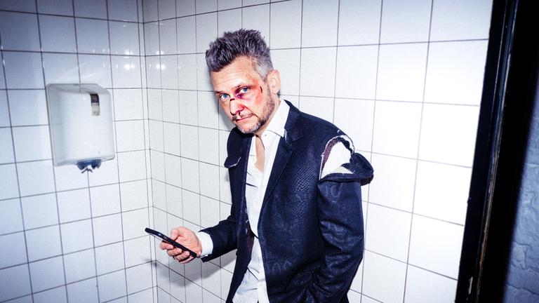 "Tobias Mann: Mann gegen Mann": Tobias Mann steht in einer öffentlichen Toilette vor dem Waschbecken, angelehnt an den Wandfliesen. Er sieht aus, als hätte er gerade eine Schlägerei gehabt, zerrissene  Klamotten, im Gesicht frische Wunden und ein blaues Auge.