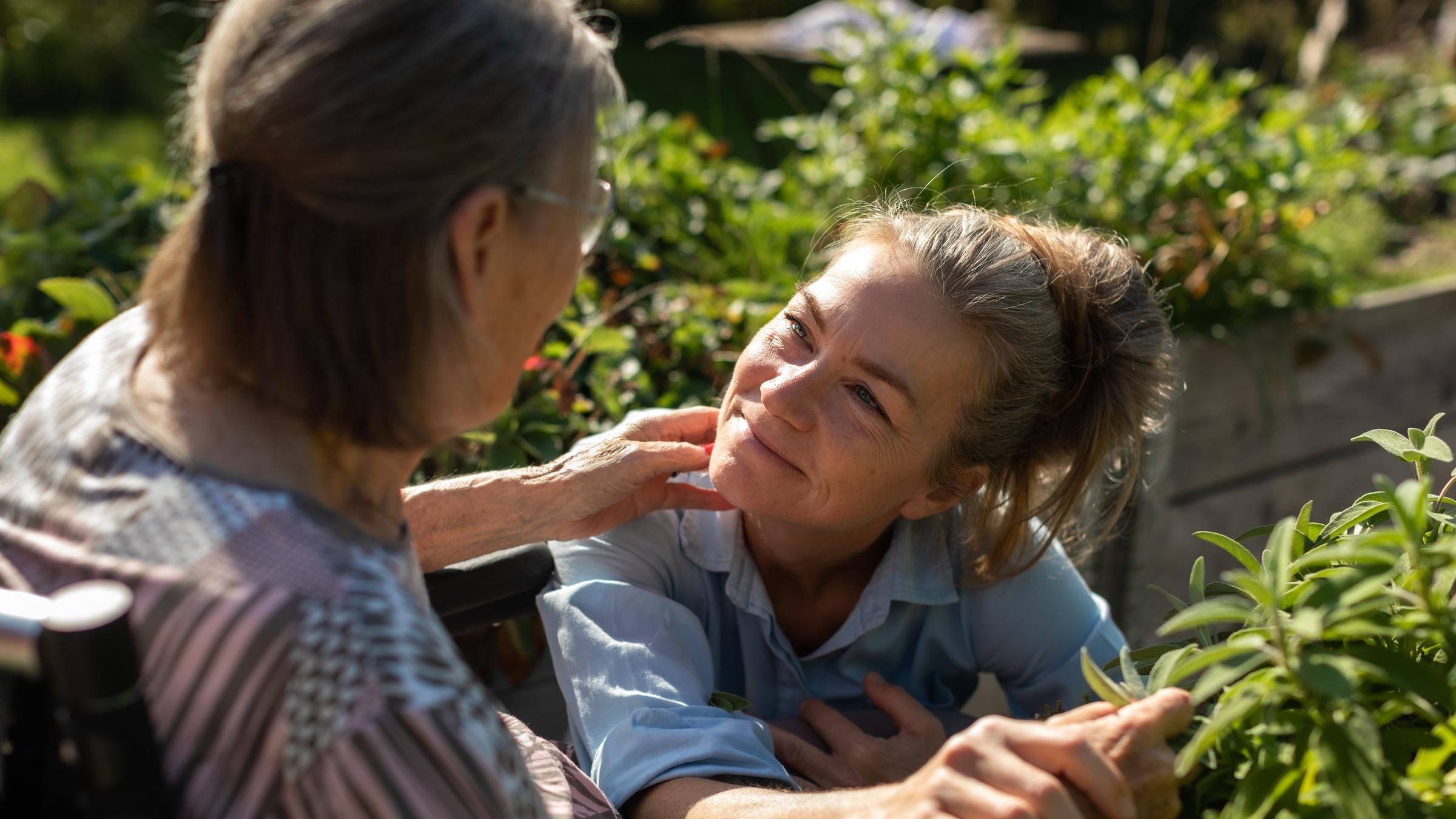 "Mitgefühl - Pflege neu denken": Alte gebrechliche Frau wird von blonder Pflegerin liebevoll gehalten.