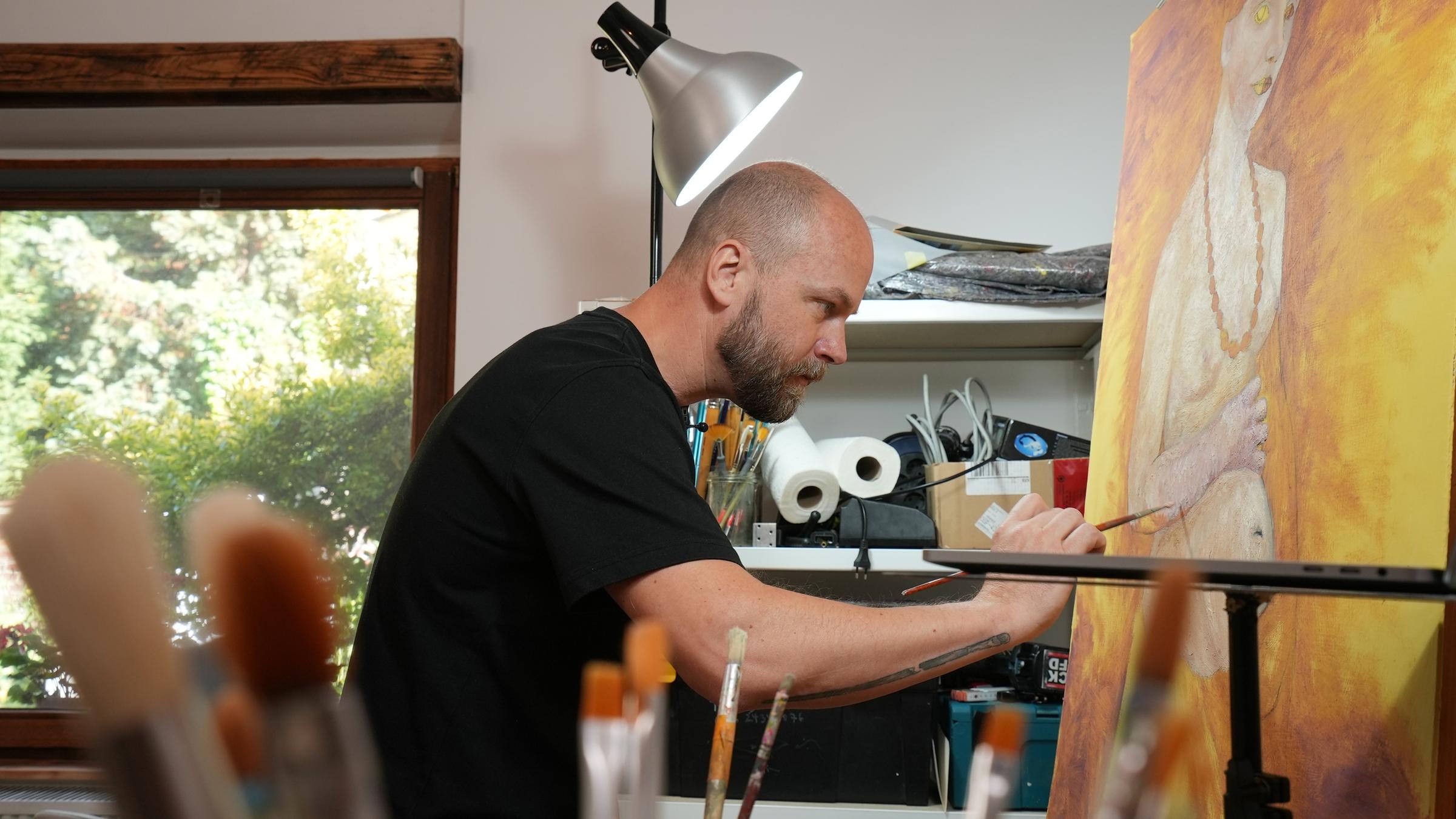 "Geheimnis der Meister: Paula Modersohn Becker": Ein Mann in schwarzem T-Shirt ist im Profil fotografiert, während er auf eine Leinwand malt. Darauf ist ein nackter, schwangerer Oberkörper erkennbar, vor einem gold-gelblichen Hintergrund.