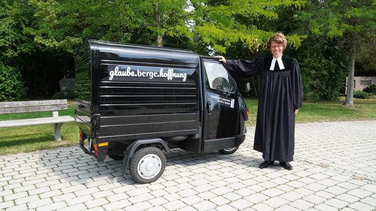 "37°: Kirche mal anders - Junge Pastor*innen im Aufbruch": Ein schwarzes Kleintransporter-Dreirad italienischer Bauart steht von links nach rechts gerichtet auf einem gepflasterten Platz. An seiner Seite prangt der Schriftzug "Glaube. Berge. Hoffnung". Rechts davon steht der junge Penzberger Pastor Philipp Roß in schwarzem Talar mit weißem Beffchen, blickt in die Kamera und stützt seine rechte Hand auf dem Fahrzeug ab. Den Hintergrund bestimmen grüne Bäume und Büsche.