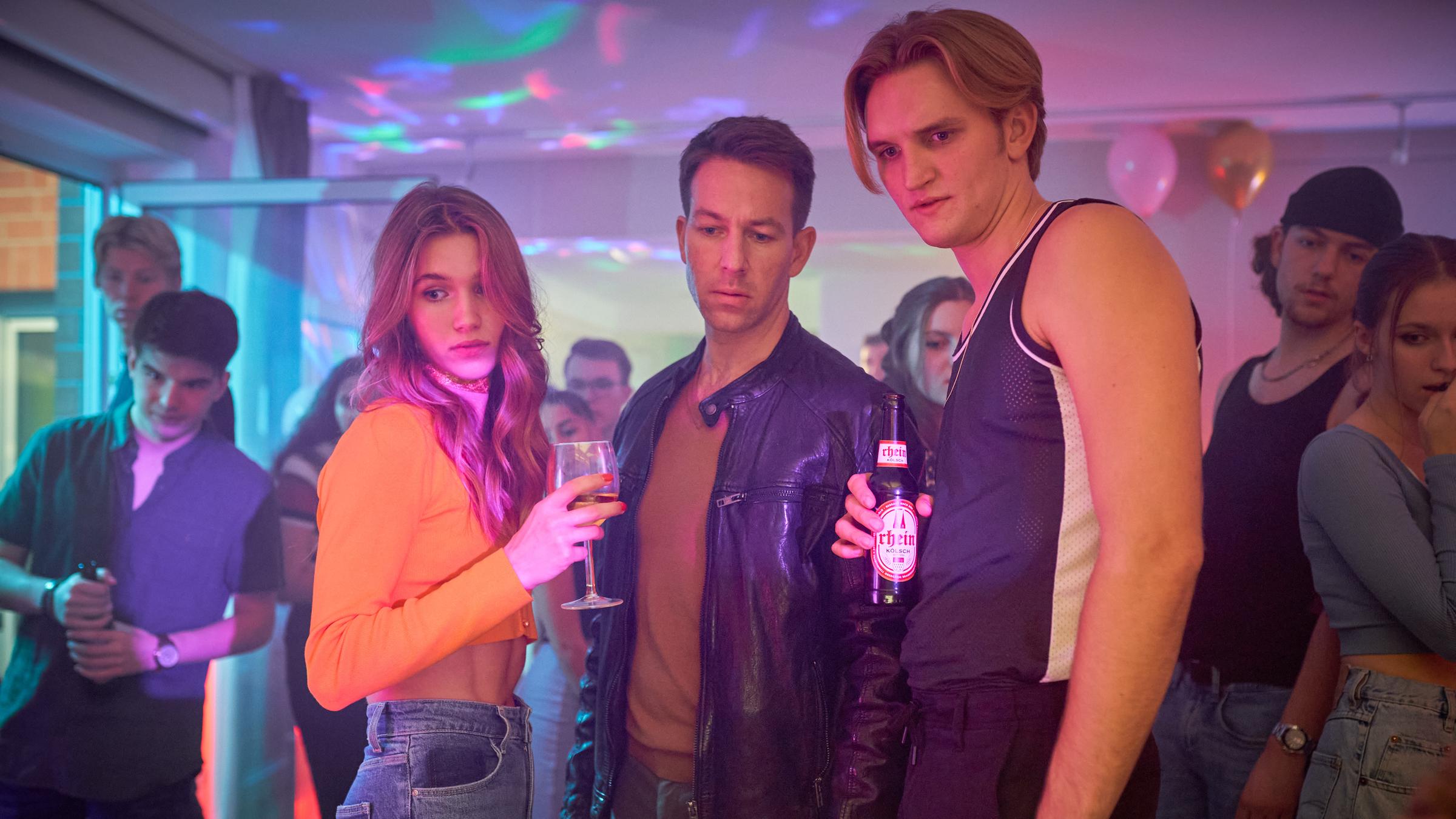  "Marie Brand und die falsche Wahrheit": Isabell Lauterbach (Anna-Lena Schwing), Trainer Victor Holtmann (Florian Anderer) und Jan Frehse (Ludger Bökelmann) stehen zusammen auf der Party und schauen auf einen gemeinsamen Blickpunkt auf dem Boden. Die Partygäste im Hintergrund (Komparsen) schauen ebenfalls dorthin. Isabell hält ein Weinglas in der Hand und Jan eine Bierflasche.