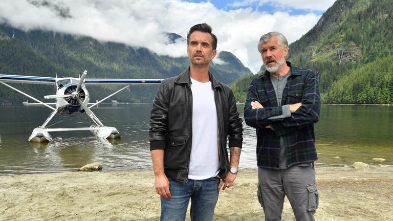 "Das Traumschiff: Vancouver": Max Parger (Florian Silbereisen) und Martin Grimm (Daniel Morgenroth) stehen am Ufer eines Sees nebeneinander. Martin hat die Arme vor der Brust verschränkt und redet mit Max. Im Hintergrund steht ein Wasserflugzeug.