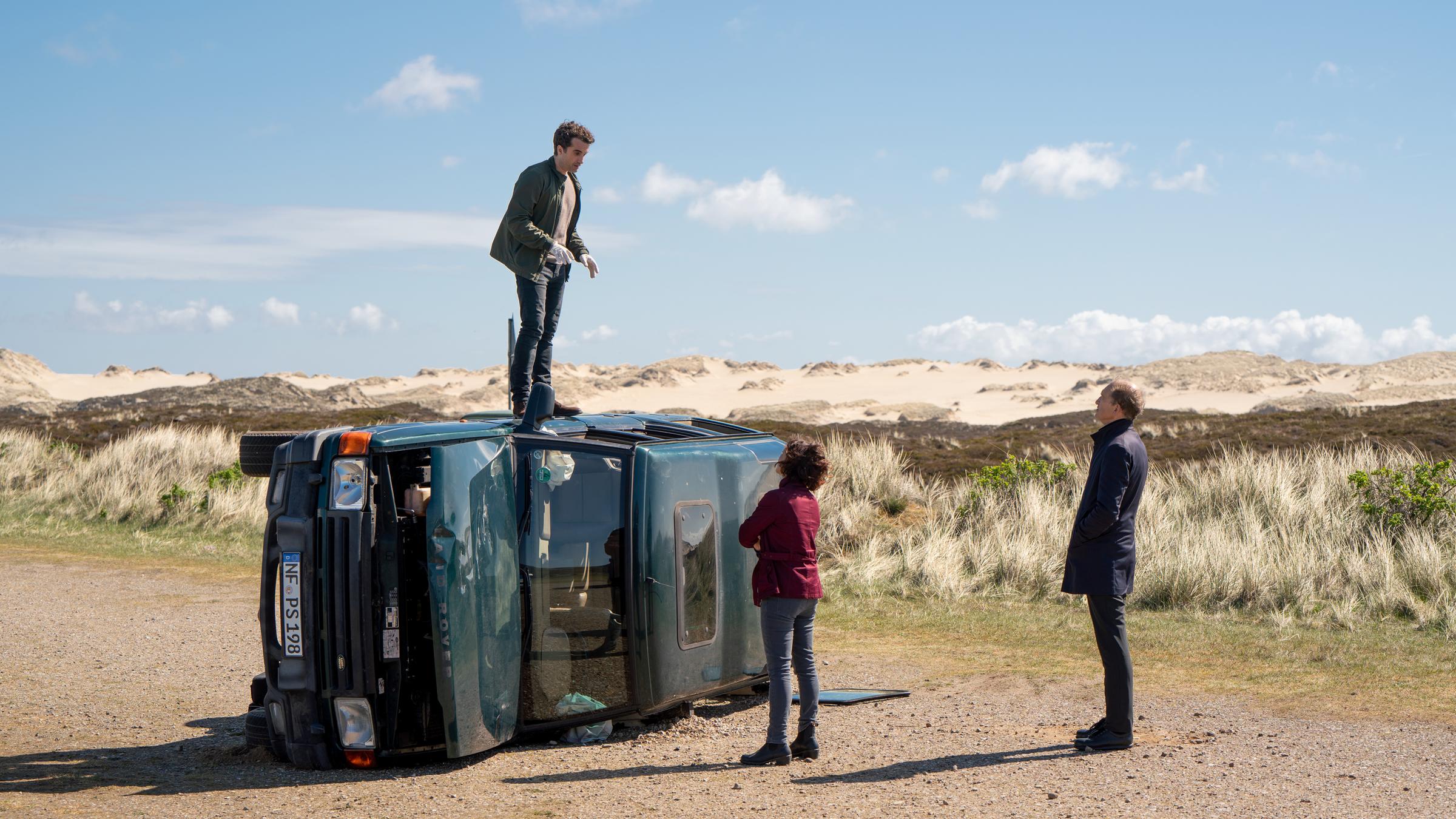 "Nord Nord Mord - Sievers und der erste Schrei": Ina Behrendsen (Julia Brendler) und Carl Sievers (Peter Heinrich Brix) stehen vor einem dunklen SUV, der demoliert auf der Seite auf einem Parkplatz in den Dünen von Sylt liegt. Hinnerk Feldmann (Oliver Wnuk) steht auf der Beifahrertür.