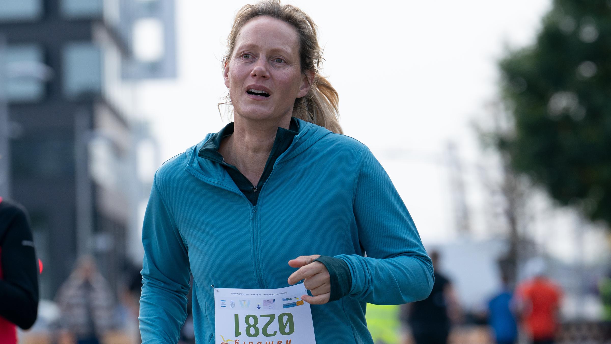 Juliane (Anna Schudt) joggt in einer blauen Laufjacke, mit einer vierstelligen Startnummer auf dem Bauch, auf einer Straße entlang.