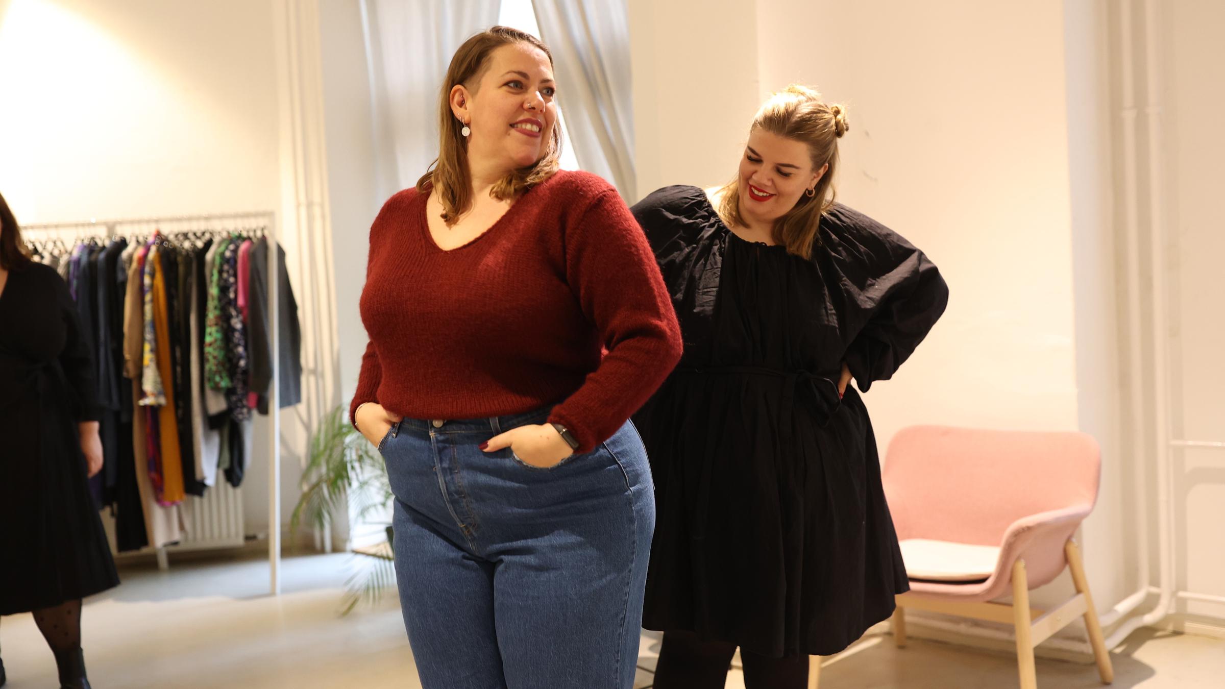 "37°: Glück kennt kein Gewicht - Mehrgewichtige und ihr neues Selbstvertrauen": Nicole, 39 Jahre alt, steht in der Bildmitte. Sie trägt Jens und einen roten Pullover, ihre dunkelblonden Haare sind halblang und am Oberkopf nach hinten gekämmt. Sie hat runde Ohrringe, ihre Lippen sind rot geschminkt. Hinter ihr steht eine junge blonde Frau, Mitte 20. Diese trägt ein elegantes schwarzes Mini-Kleid mit Puffärmeln. Sie guckt auf die andere Frau, die offensichtlich die Kleidungsstücke probiert und sich im Spiegel betrachtet, der jedoch außerhalb des Bildes ist.