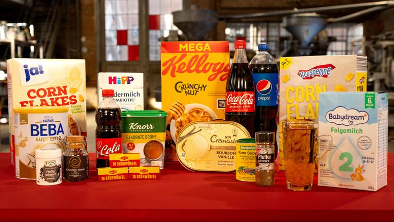 "ZDFbesseresser: Tricks der Lebensmittelindustrie weltweit - Sebastian Lege deckt auf": Verschiedene Produkte wie Cornflakes, Cola, Eis und Baby-Milchpulver stehen auf einem Tisch.