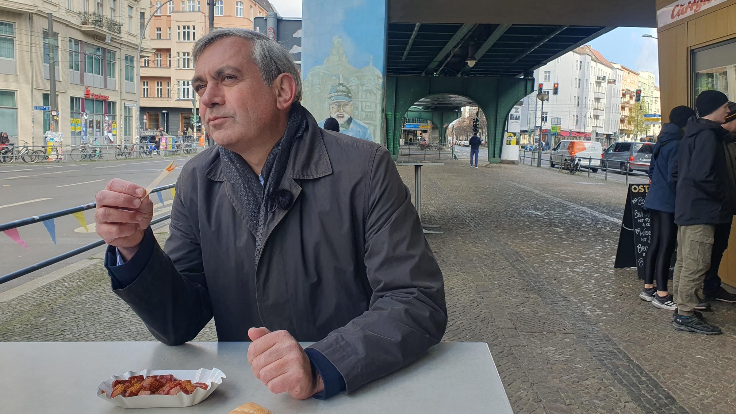"Kaminer Inside: Wie isst Deutschland?": Kaminer steht unter einem U-Bahn-Bogen an einem Stehtisch und verspeist genüsslich eine Currywurst. Am Torbogen hinter ihm als Graffiti ein preußischer Offizier mit Pickelhaube.