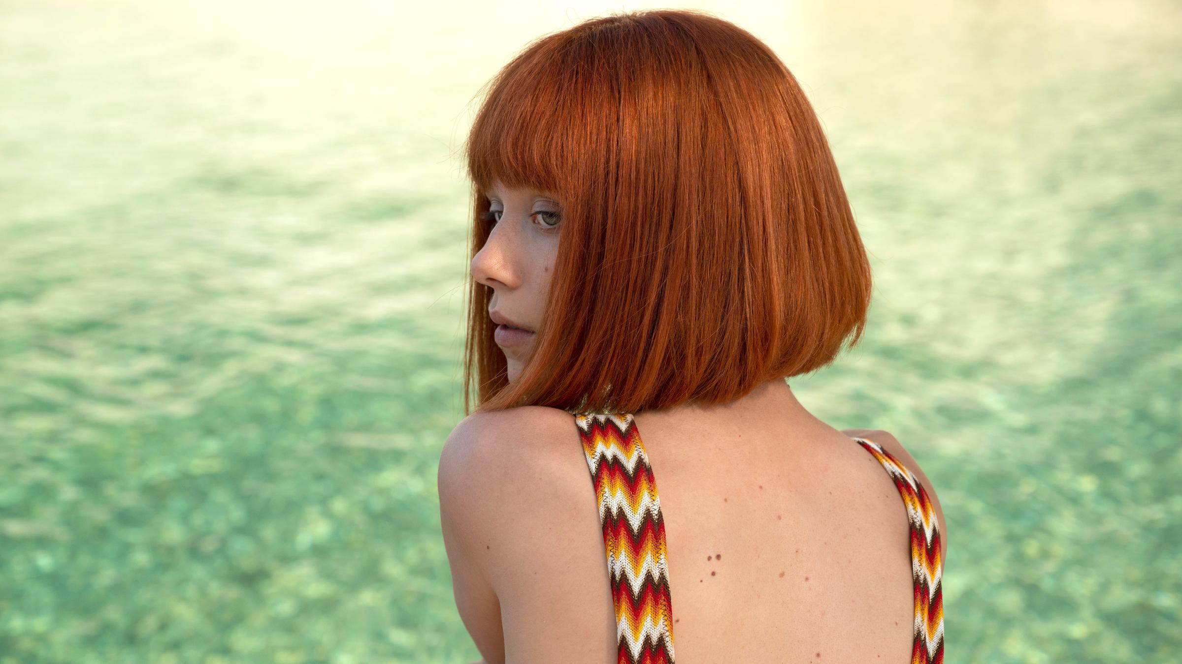 "Das Mädchen und die Nacht (1)": Vinca (Ivanna Sakhno) sitzt am Wasser. Sie trägt einen bunt gemusterten Badeanzug.