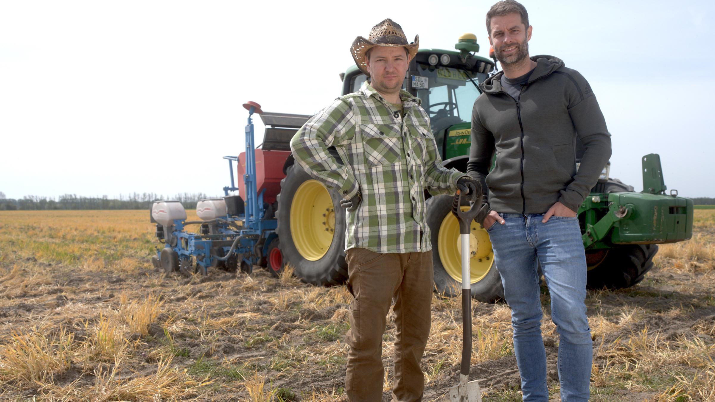"ZDFzeit: Trocknet Deutschland aus? Gefahren, Fakten, Lösungen": Vor einem Traktor stehen Landwirt Michel Allmrodt und Kai Kupferschmidt auf einem Acker. Michel Allmrodt trägt ein kariertes Hemd, einen Hut und stützt sich auf einen Spaten, Kai Kupferschmidt hat beide Hände in den Hosentaschen.