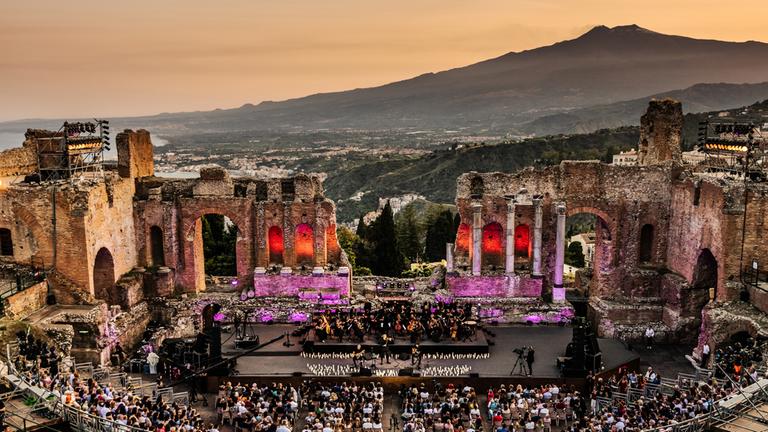 "David Garrett auf Sizilien": Im Vordergrund liegt das beleuchtete antike Theater von Taormina mit besetzten Zuschauerrängen, auf der Bühne sind David Garrett und das Orchester. Im Hintergrund, im Abendlicht, die Silhouette des Ätnas und die sizilianische Mittelmeerküste.