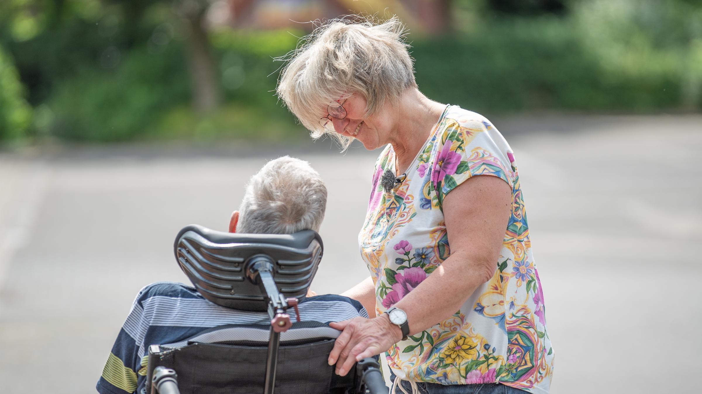 "37 Grad: Ich entscheide für Dich - Wenn Angehörige zu Betreuern werden": Hinterkopf eines Mannes, der in einem Rollstuhlsitzt, daneben steht eine Frau und beugt sich leicht zu ihm vor. Ihre Hand liegt dabei auf der Lehne des Rollstuhls.