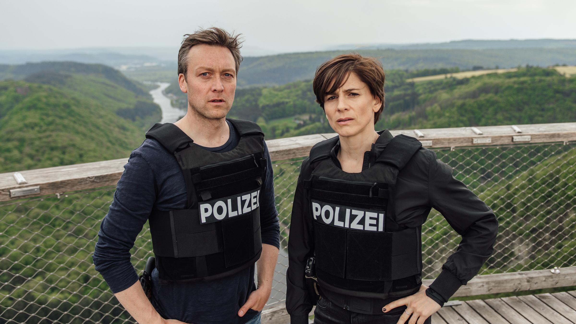 „In Wahrheit – Blind vor Liebe“: Das Ermittlerteam Judith Mohn (Christina Hecke) und Freddy Breyer (Robin Sondermann) steht auf einer Aussichtsplattform mit Ausblick auf die Saarschleife. Sie tragen beide Schutzwesten.