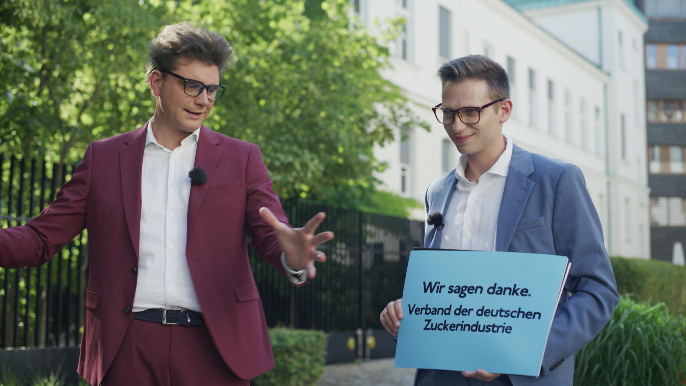 "heute-show spezial": Lutz van der Horst und Fabian Köster stehen nebeneinander im Freien, Fabian Köster hält ein Plakat mit der Aufschrift "Wir sagen danke. Verband der deutschen Zuckerindustrie".