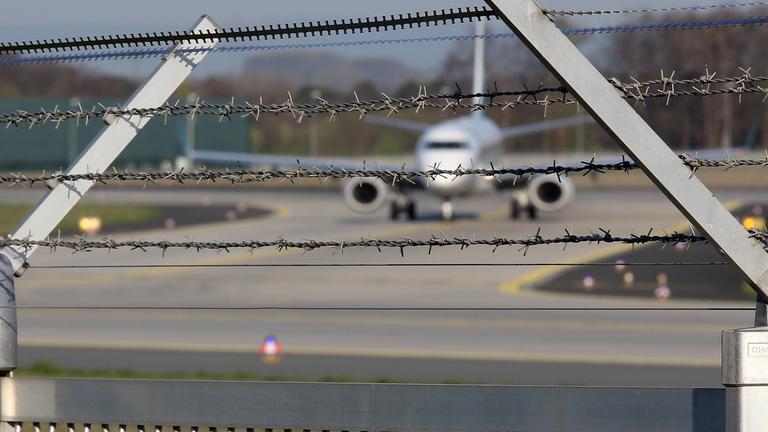 "#WTF - Blackbox Abschiebung": Symbolbild: Blick durch einen Stacheldrahtzaun auf ein Flugzeug, das auf dem Rollfeld steht.