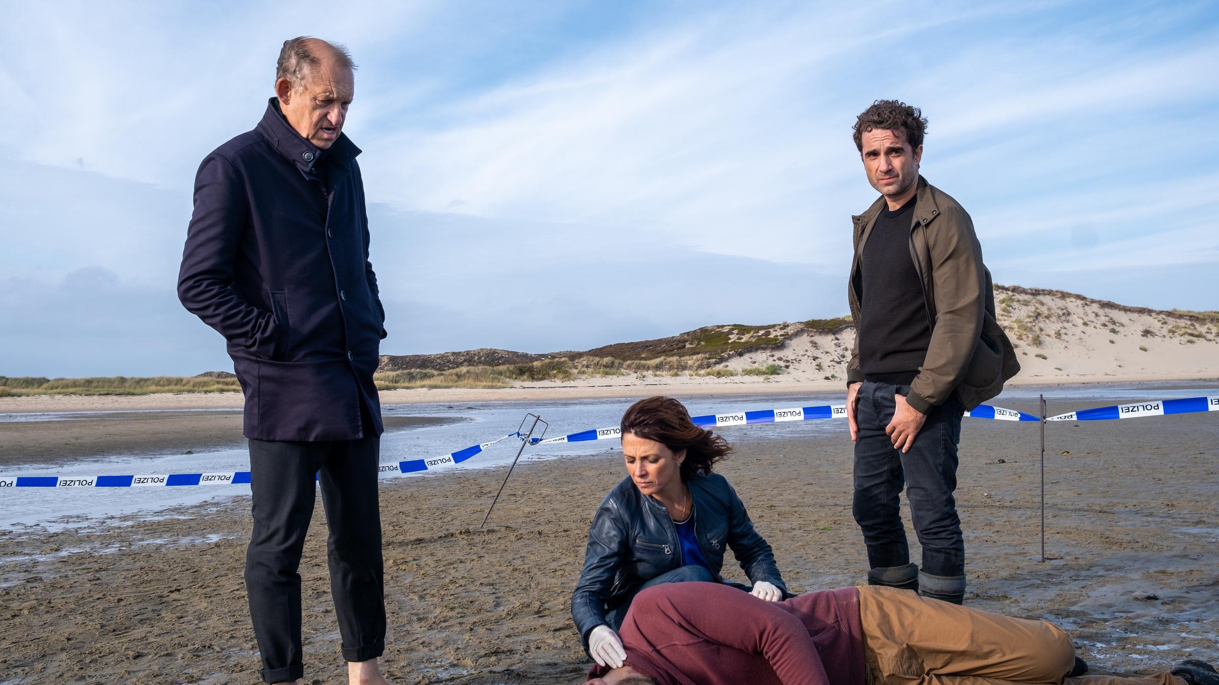 "Nord Nord Mord - Sievers und der Traum vom Fliegen": Carl Sievers (Peter Heinrich Brix) steht neben Ina Behrendsen (Julia Brendler), die sich gerade zur Leiche von Pablo Hernandez (Iskander Madjitov) herunterkniet, um dessen Puls zu fühlen. Neben Ina steht Hinnerk Feldmann (Oliver Wnuk), der die Hände in seinen Gürtelschlaufen eingehakt hat. Die Kommissare befinden sich im Watt und der Fundort ist mit einem blau-weißen Band der Polizei abgesperrt.