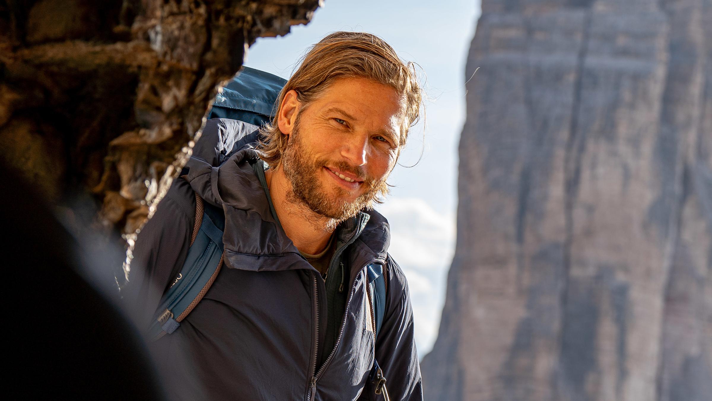 „Sebastian Ströbel – Meine Alpen, Menschen der Berge“: Sebastian Ströbel in dunkler Jacke und blauem Rucksack am Eingang des Weltkriegsstollens in Paternkofen in den Dolomiten.
