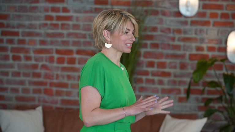 "Buchstäblich leben": Sabine Heinrich steht in Grün gekleidet vor einer Backsteinwand und spricht zu nicht sichtbaren Anwesenden.
