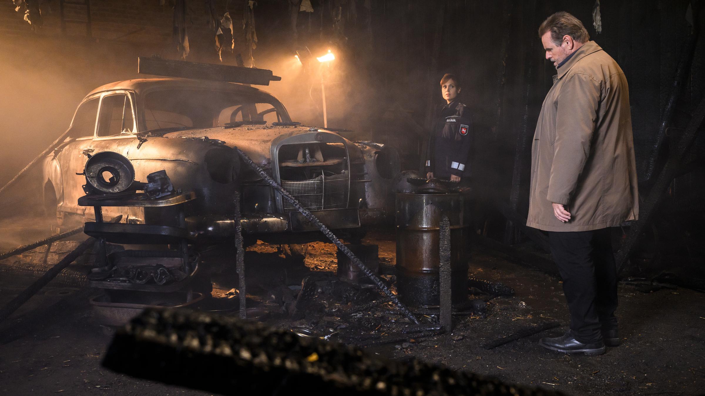 "Friesland – Feuerteufel": Süher Özlügül (Sophie Dal) und Jan Brockhorst (Felix Vörtler) stehen in einer Scheune vor einem abgebrannten Oldtimer. Süher blickt zu Brockhorst, der den Boden des Tatorts im Visier hat.