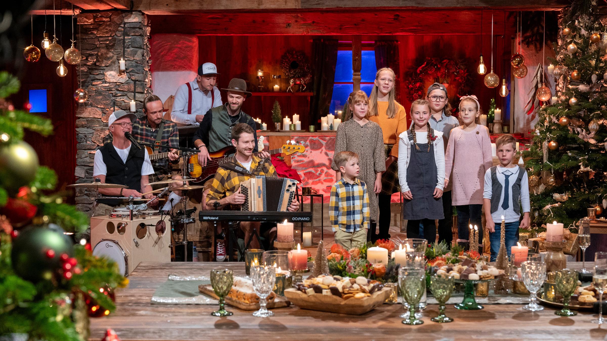 "Heiligabend mit Carmen Nebel": Die Hüttenband Tschentig und die Ellmauer Kinder stehen in weihnachtlicher Kulisse auf der Bühne. Um sie herum gibt es weihnachtliche Dekoration, Weihnachtsbäume, brennende Kerzen und einen geschmückten Tisch.