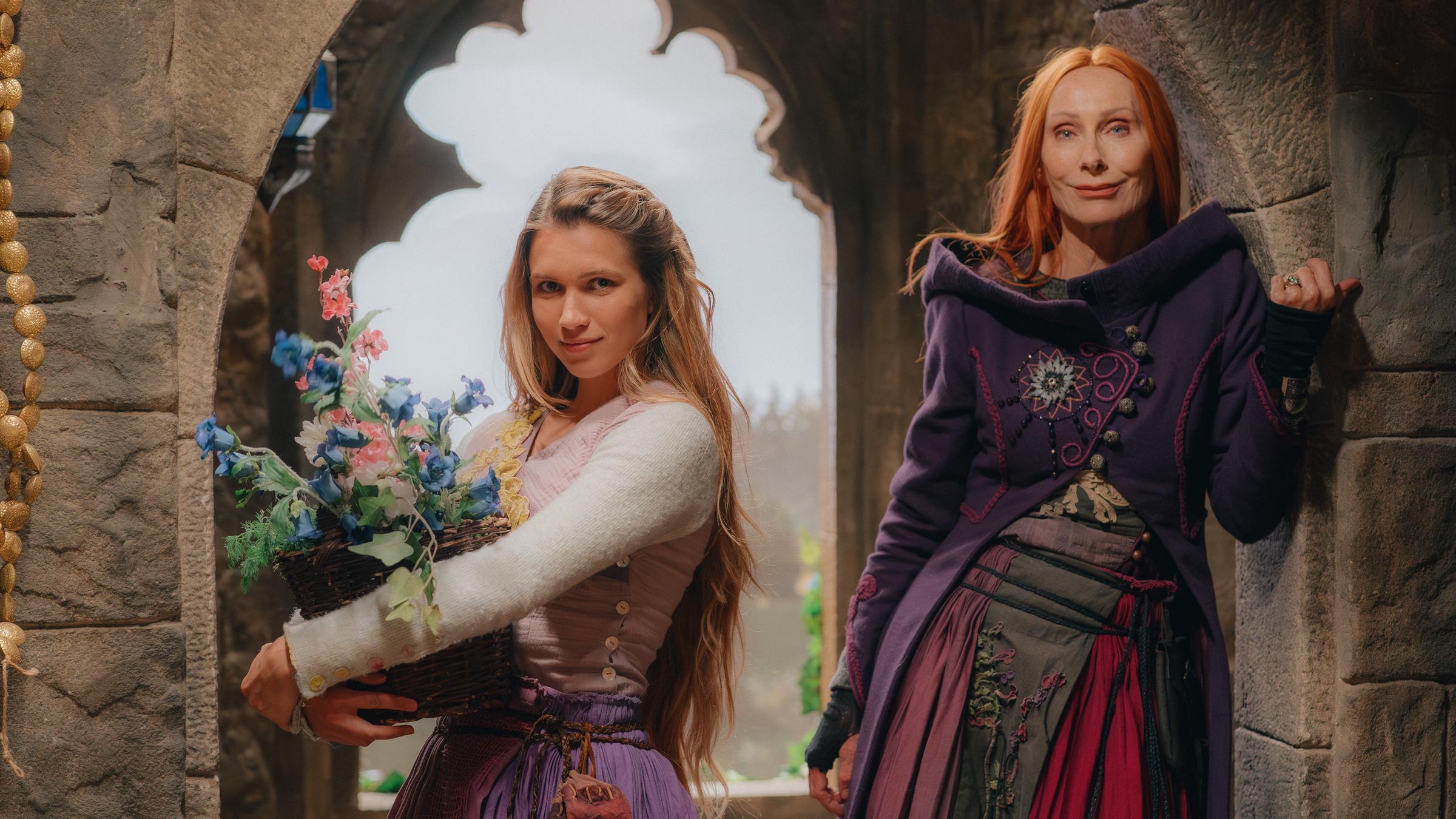 "Rapunzel und die Rückkehr der Falken": Rapunzel (Anna-Lena Schwing) mit einem Blumenkorb und Zauberin Eleonor (Andrea Sawatzki) stehen in einem mittelalterlichen Turmzimmer vor einem Fenster. Beide schauen lächelnd zur Kamera.