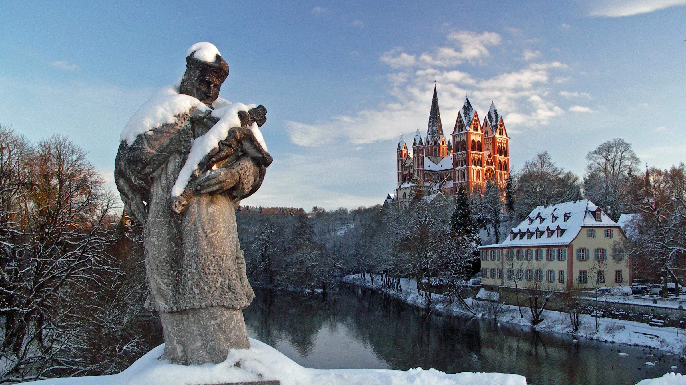 "Katholischer Weihnachtsgottesdienst - Ehrenwort!":  In einer Winterlandschaft steht im Vordergrund die Statue des Heiligen Johann von Nepomuk, dem Schutzpatron der Brücken, auf einer Brücke über die Lahn. Dahinter liegt der Limburger Dom im Sonnenschein.