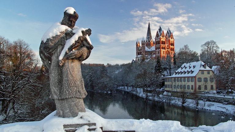 "Katholischer Weihnachtsgottesdienst - Ehrenwort!":  In einer Winterlandschaft steht im Vordergrund die Statue des Heiligen Johann von Nepomuk, dem Schutzpatron der Brücken, auf einer Brücke über die Lahn. Dahinter liegt der Limburger Dom im Sonnenschein.