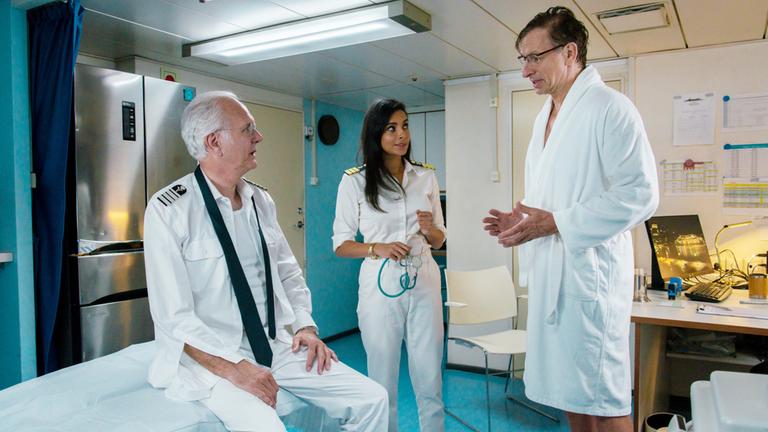 "Das Traumschiff - Utah": Im Arztzimmer des Schiffs: Mit geöffnetem Hemd und Krawatte sitzt Oskar Schifferle (Harald Schmidt) auf der Untersuchungsliege. Er blickt irritiert zu Jens Kessler (Ingolf Lück), der mit verzweifelten Blick und gestikulierend im Raum steht.  Zwischen den Männern steht Dr. Jessica Delgado (Collien Ulmen Fernandes). Sie hält ein Stethoskop in den Händen und blickt fragend Jens Kessler an.