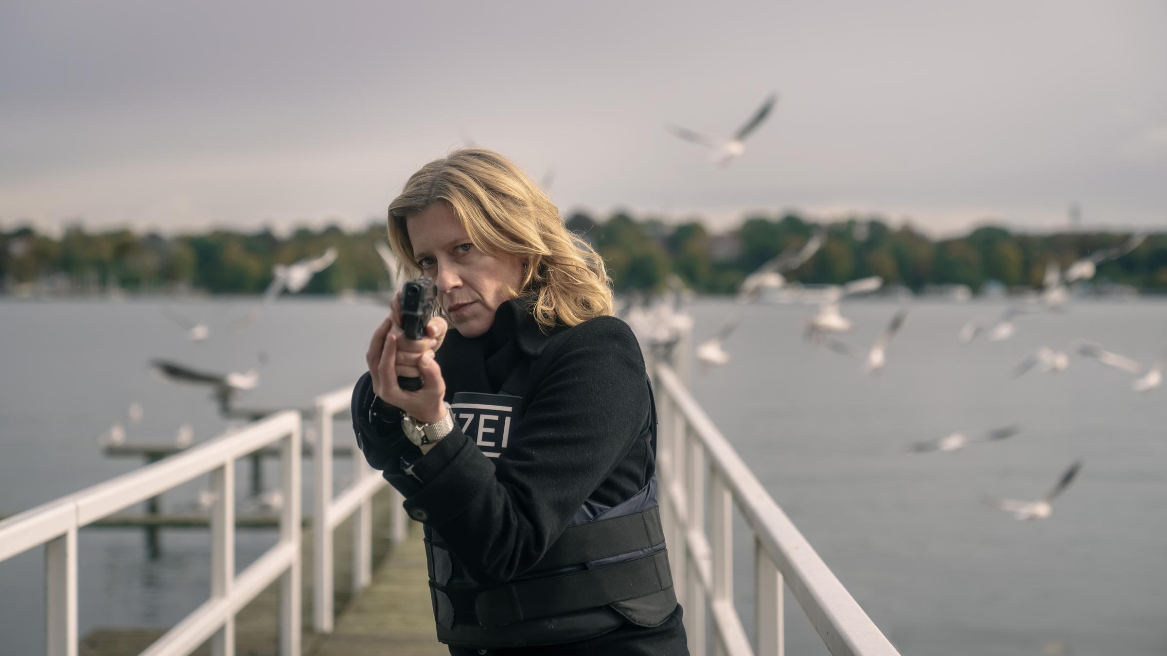 "Kolleginnen - Abgetaucht": Irene Gaup (Caroline Peters) zielt frontal mit einer Pistole, während sind auf einem Steg steht und hinter ihr Seevögel die über das Wasser fliegen.