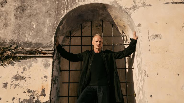 Sting: My Songs - Live at Château de Chambord(3sat): Sting posiert, schwarz gekleidet, in einem alten Mauerportal und berührt mit ausgebreiteten Armen die Innenwände.