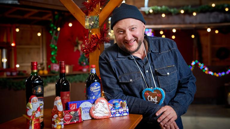 "besseresser: Die Tricks in Glühwein, Lebkuchen & Co. - Sebastian Lege deckt auf": Produktentwickler Sebastian Lege steht an einem Weihnachtsmarkt-Stand, um seinen Hals trägt er ein Lebkuchenherz mit der Aufschrift "Chef", neben ihm auf dem Tisch sind Glühweinflaschen, Schoko-Nikoläuse und Dominosteine angeordnet.